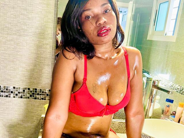 Makena69 with shiny skin, wearing a red bra, has her tits out, showing off her shiny, wet chest, and red lipstick, sitting in a bathroom, looking at the camera.