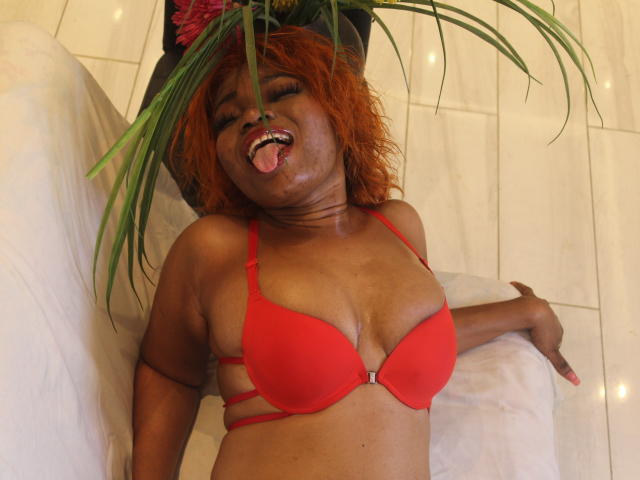 Makena69 with red bra, orange hair, tongue out, eyes closed, leaning on white pillow, green plant leaves in hat, brown skin, medium tits, strappy bra, pink nails, smiling.