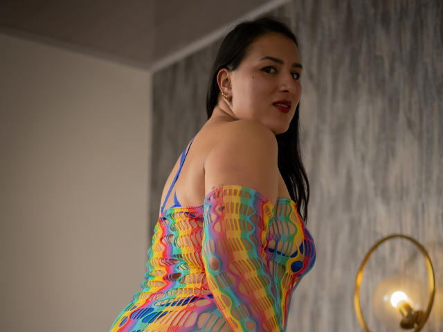 KailanyCooper with dark hair, red lipstick, and colorful, see-through, wavy-patterned dress, showing her back and looking over her shoulder, in a room with a lamp.
