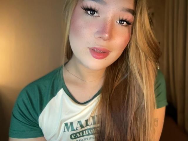 MistressAmericana has long blonde hair, green eyes, and pink lips with a slight smile. She's wearing a green and white raglan shirt, and her freckled face is cute.