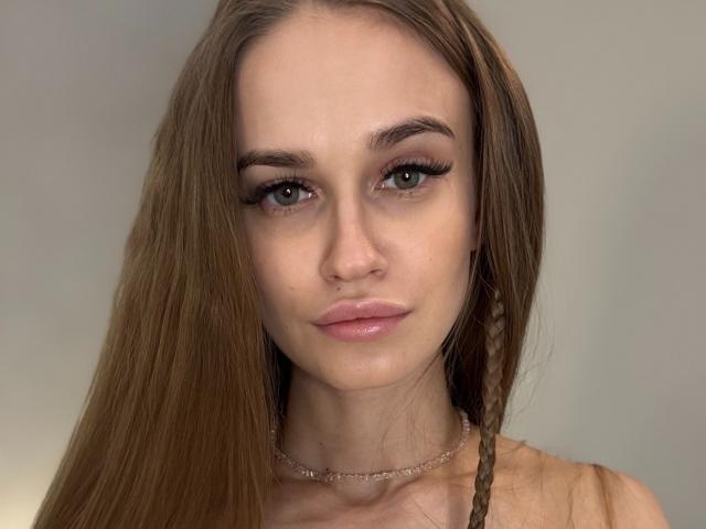 AmmiSun has long brown hair, green eyes, and glossy pink lips. She's wearing a thin necklace and has a small braid on the right side of her face.
