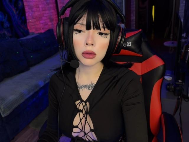 AmeliaAley with black bob, wearing headphones, red-black chair, black lace-up top, showing her tits, has tattoos, pink lipstick, and a slightly bored expression in a dimly lit room.