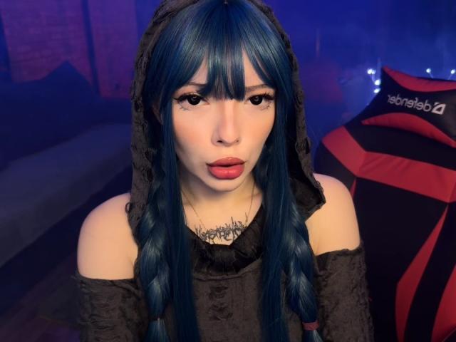 AmeliaAley with blue braided hair, black lace hood, off-shoulder top, red lipstick, tattoo on neck, and pouty lips. She's sitting in front of a red and black gaming chair.
