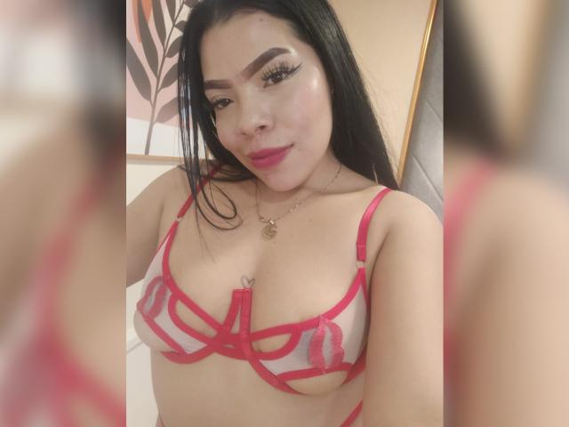AdrianaPaul with black hair, pink lipstick, and heavy eyeliner, wearing a red strappy lingerie set, showing her tits, smiling, necklace, and a mirror reflection in the background.