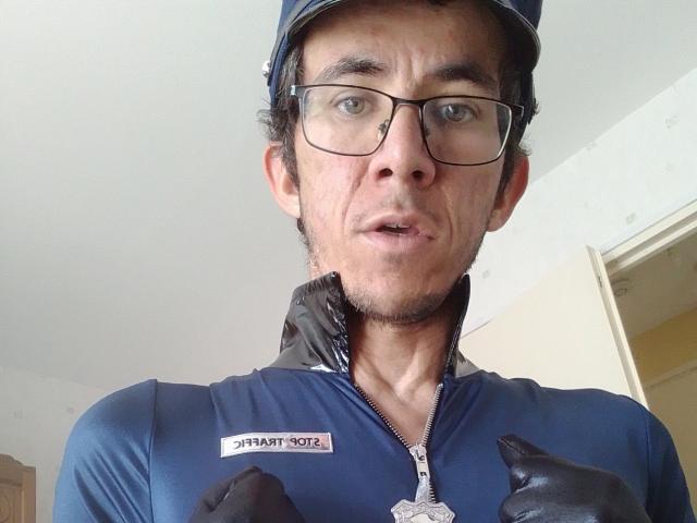Bricebd, wearing a blue cycling jersey, black gloves, and glasses, with a slightly open zipper, looks into the camera. He has a short beard and is wearing a cap.