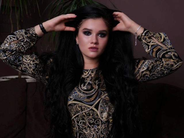 KristiTopxx-hot tem cabelos longos e pretos, um vestido estampado com desenhos em espiral, e está olhando para a câmera com as mãos no cabelo. Ao fundo, há uma parede marrom e um sofá marrom escuro..