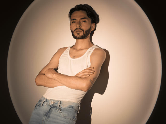 JakeMagnus with arms crossed, wearing a white tank top and blue jeans, standing against a lit, circular, shadowed background, has a beard, short dark hair, and a tattoo.