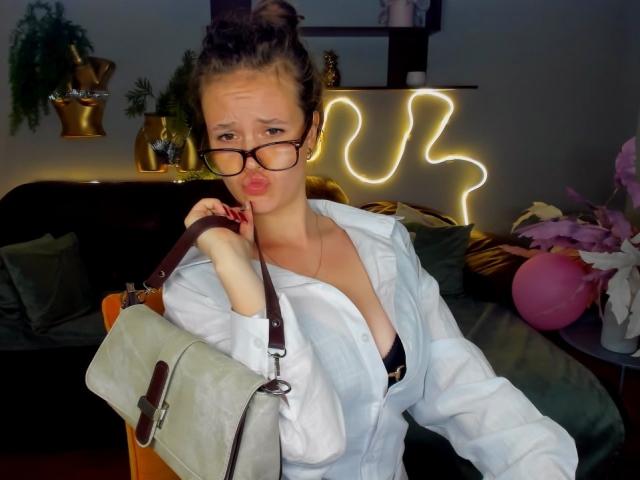 BeatriceKeat with glasses, pouty lips, white shirt, black bra, small tits, grey bag, brown hair bun, neon wall light, green couch, pink flowers, and a pillow in the background.