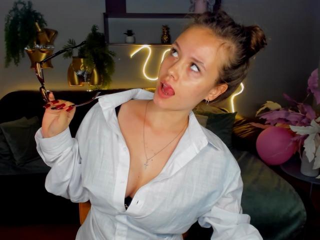 BeatriceKeat with curly brown hair in buns, white button-up shirt, black bra, holding a pipe, tongue out, looking to the side, in a dimly lit room.