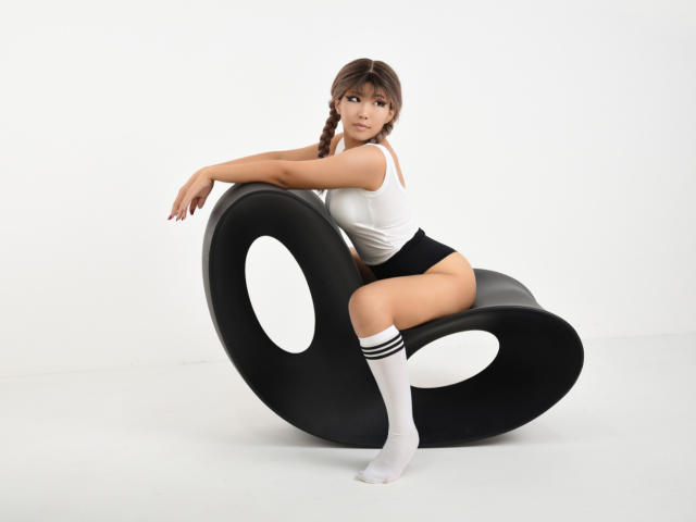WendyFire with brown pigtails, white tank top, black high-waisted shorts, and white socks with black stripes, sitting on a black, curved chair, looking at camera.