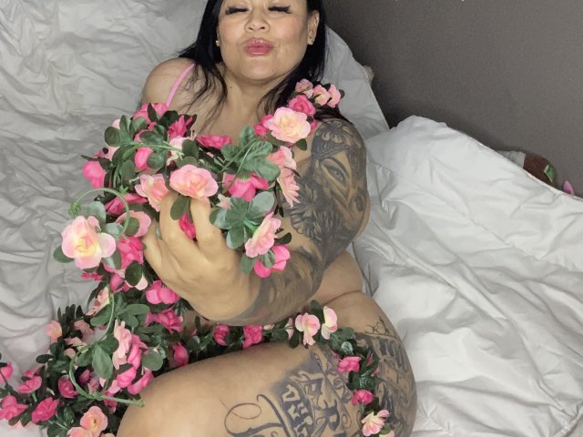 KittyHotLady69 with pink roses, tattoos covering her arms and legs, puffy white bed, sitting, holding roses to her chest, puckering lips, dark hair, pink lingerie, sexy.