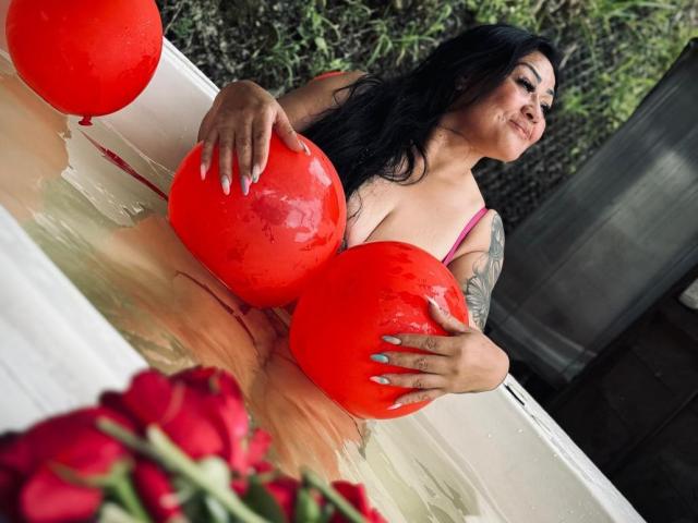KittyHotLady69 avec ses cheveux noirs mouillés, un soutien-gorge rose, tenant deux ballons rouges au-dessus de ses seins, dans une baignoire blanche remplie de pétales de roses rouges, souriante, avec un tatouage sur le bras droit..