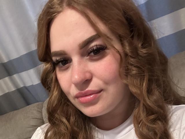 ChattieIssy has curly brown hair, long eyelashes, pink glossy lips, and a cute, slightly flushed face. She's wearing a white shirt, looking softly at the camera.