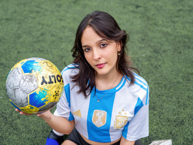 LanaKlaire, con capelli castano scuro, indossa una maglia da calcio blu e bianca, tiene in mano un pallone da calcio giallo e blu sporco, è seduta sull'erba verde e guarda verso l'alto con il rossetto rosa..