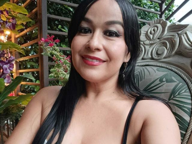 TamaraFoxy has long black hair, red lipstick, and is smiling at the camera with her tits visible in a low-cut top, wearing hoop earrings. She's sitting outdoors.