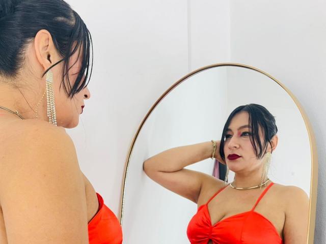 EmaFoxy with dark hair in a red dress, reflecting in a gold mirror, showing her tits, red lipstick, and gold earrings, with a hand in her hair.