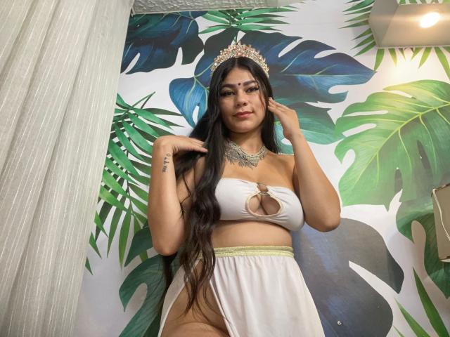 KeraniMarala, met lang zwart haar, draagt een witte top met uitsparingen, waardoor haar borsten zichtbaar zijn, en een witte rok. Ze heeft een sierlijke ketting en een kroon, en haar handen zijn in de buurt van haar gezicht..