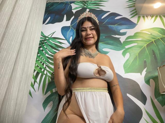 KeraniMarala with long black hair, wearing a white top with a keyhole cutout, exposing her tits, and a sheer white skirt, has a crown, tattoos, and a necklace.