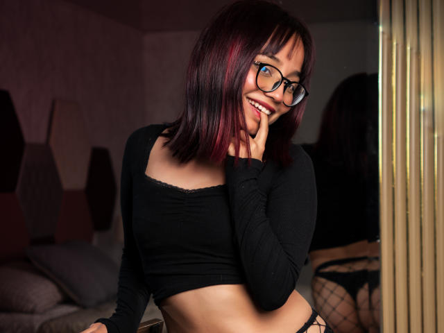 LeilaIvanova with red and black shoulder-length hair, glasses, smiling, black crop top, black panties, finger on cheek, mirror reflection, bedroom, sexy, cute, attractive, small tits, confident.