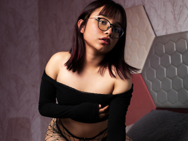 LeilaIvanova with glasses, black off-shoulder top, fishnet pants, and exposed tits, leaning forward, looking at camera, with short dark red hair, in a dimly lit room.