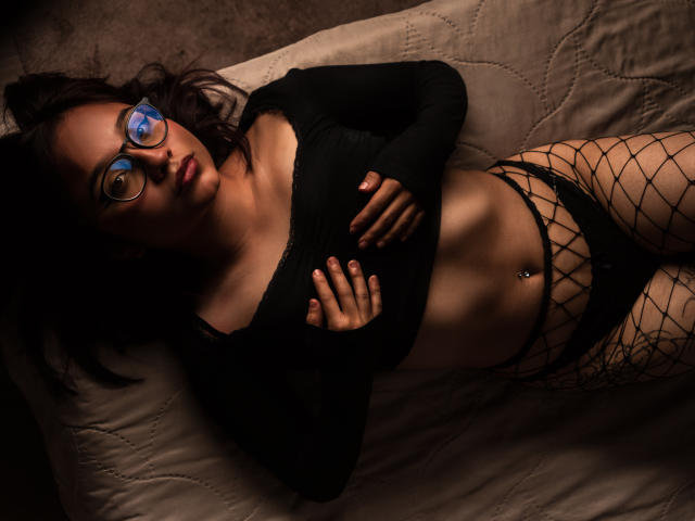 LeilaIvanova lies on a bed, wearing glasses, black long-sleeve crop top, fishnet pants, and black panties, exposing her toned abs and belly button piercing. Her hands touch her chest.