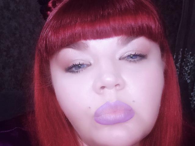 RedBustyX with bright red hair, pale skin, and purple lipstick, has a pouty face and small moles on her nose. Her blue eyes are slightly open.