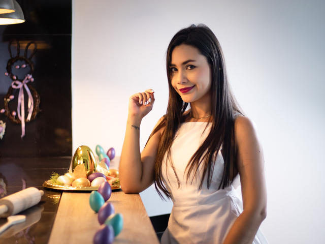 SophieJones69, con cabello castaño largo, vestida con un vestido blanco, sosteniendo un chocolate, de pie junto a una mesa con huevos y hojas moradas y verdes, sonriendo. Sus pechos son pequeños, su trasero es lindo..
