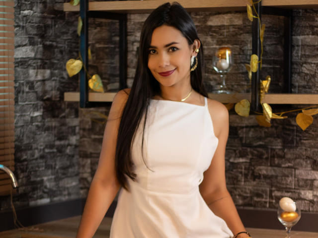 SophieJones69 with long black hair, wearing a white sleeveless dress, smiling at the camera, showing her tits, in a dark stone wall setting, holding a wine glass.