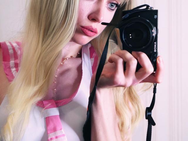 Kirana, avec ses cheveux blonds, portant un haut rose et blanc, tient une caméra Sony noire près de son visage, montrant ses lèvres roses et pulpeuses ainsi qu'un collier de perles. Le bracelet de la caméra est visible..