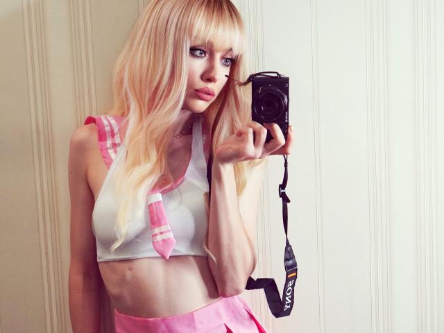 Kirana, met blond haar, gekleed in een roze en witte schoolmeisjesoutfit, houdt een camera vast en laat haar kleine borsten zien in een witte crop top, een roze rok en een camerastrap..