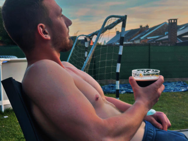 DeltaOchoX, shirtless, muscular, holding beer, sitting on chair, grassy backyard, soccer goal, sunset, right profile, brown hair, beard, blue jeans, nipple piercing, relaxed, casual.