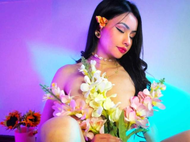 KattRouse with black hair, orange flower in her hair, pink and white flowers covering her bare tits, wearing a gold necklace, sitting, pink and blue lighting.