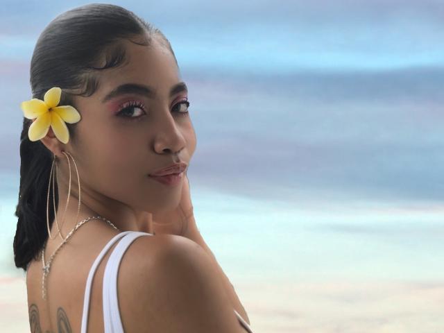 AvaRouge with a yellow plumeria in her black hair, hoop earrings, and white tank top, looking over her shoulder, showing her bare back and small tattoo.