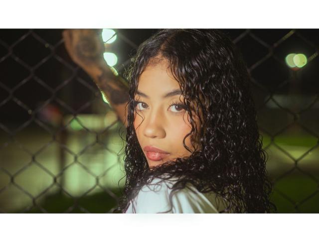 AvaRouge with curly black hair, looking over her shoulder, pouty lips, and brown eyes, standing in front of a chain-link fence, with a tattooed arm in the background.