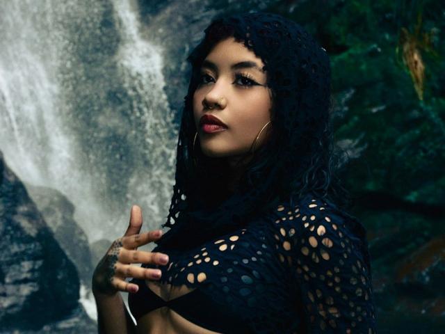 AvaRouge with curly black hair, wearing a black crochet top, showing her tits, has a nose ring, hoop earrings, and red lipstick, standing in front of a waterfall.