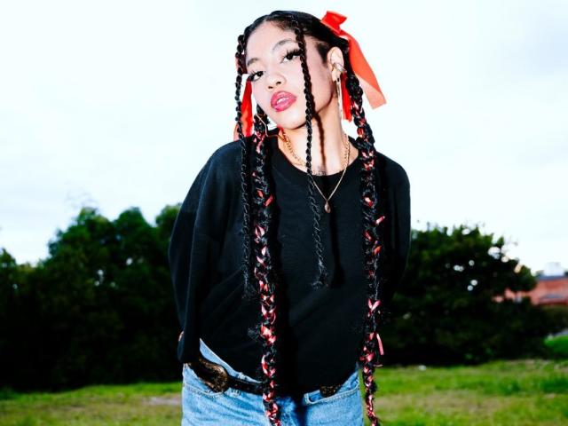 AvaRouge with long braided black and red hair, red bow, black sweater, blue jeans, hands in pockets, standing outside, looking at camera, slightly tilted head, subtle smile.
