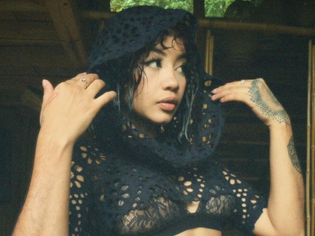 AvaRouge with wet black lace top, holding black crocheted hood, showing tattoos on right hand, looking side, nipples visible through lace, natural light, forest background.