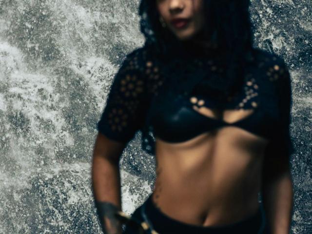 AvaRouge with dark curly hair, black lace top, and black leather bra, showing her toned abs and small tits, wearing black pants and a black glove.