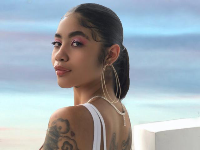 AvaRouge with dark hair in a ponytail, wearing hoop earrings and a white top, showing tattoos on her right arm, looking over her shoulder with pink eyeshadow.