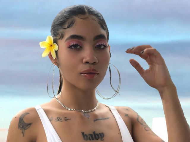 AvaRouge with dark hair, yellow flower, pink eyeliner, hoop earrings, white tank top, tattoos, and a chain necklace, showing off her tits and a hand. Beach background.