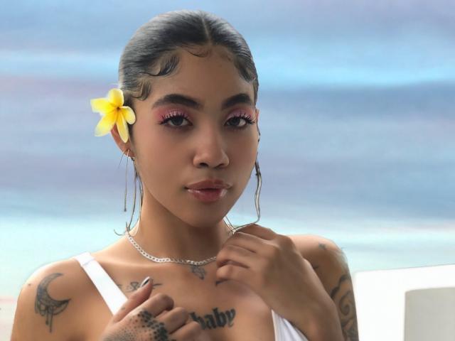 AvaRouge with tan skin, dark hair, pink eyeshadow, and yellow flower in hair, wearing white top, touching chest with tattoos, small tits, and silver necklace.