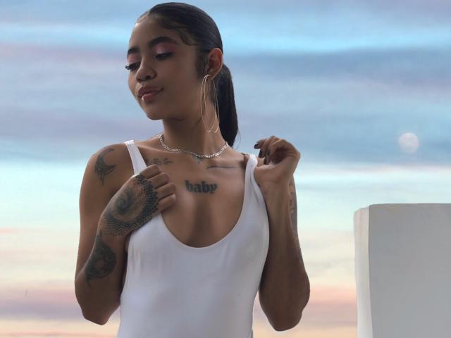 AvaRouge with dark hair, white tank top, tattoos, hoop earrings, and a necklace, holding her shirt, showing her small tits and a chest tattoo. Sunset background.