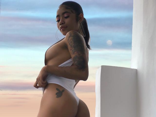 AvaRouge with a tattoo on her left hip, wearing a white thong that reveals her ass and sideboob, has tattoos on her arm, and large hoop earrings.