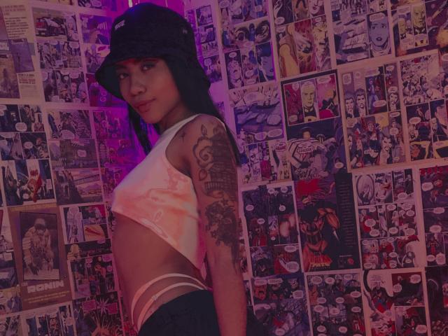 AvaRouge with tattoos, wearing a black bucket hat and white tie-dye crop top, showing her ass in strappy underwear, standing against a wall of comic book posters.