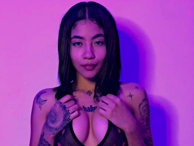 AvaRouge with dark hair, tattoos on arms and chest, holding her tits in a black mesh top, purple and pink lighting, looking at the camera with a slight pout.