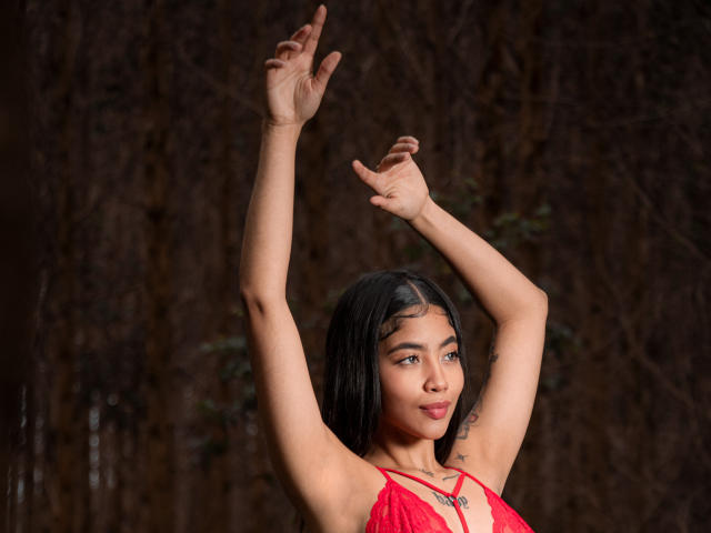 AvaRouge with tan skin and black hair, arms raised, wearing a red lace top, showing her tits, looking to the side with a slight smile.