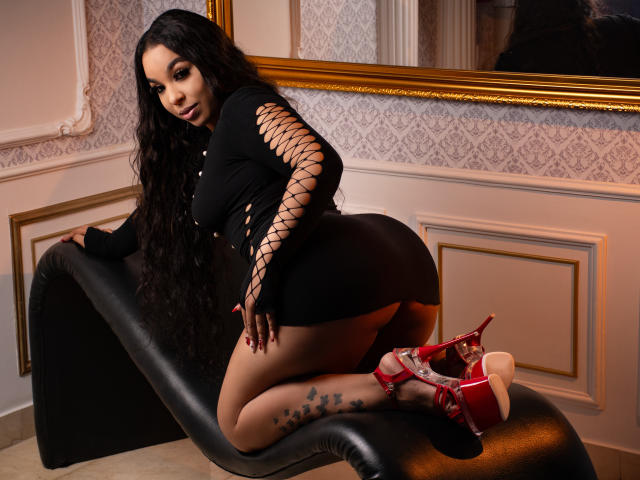 EileenBenzon kneels on a black chaise, wearing a black lace-up top and red high heels, with her big ass and star tattoos visible, long black hair flowing.