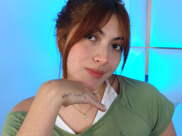 SpencerLe, con cabello castaño, camisa verde, camiseta blanca debajo, tatuaje en la mano que dice "Te amo" con un corazón, fondo azul, con el mentón apoyado en la mano, una sonrisa sutil, piel clara, linda..