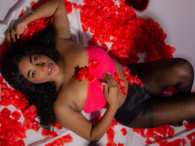 CharloteBrown with curly black hair, lying on red rose petals, wearing a pink strapless top, black lace panties, and black stockings, looking at the camera with a sexy smile.