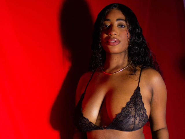 Charlote Brown, met donker krullend haar, draagt een zwarte kanten BH en laat haar borsten zien, met een rode achtergrond, kijkend naar de camera met een lichte glimlach..